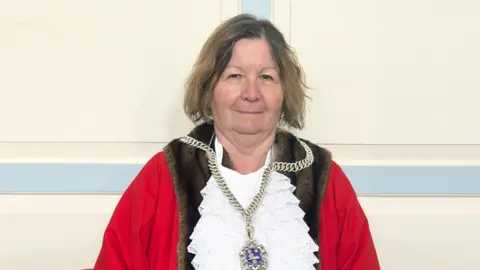 A woman with shoulder-length brown hair. She is dressed in mayoral robes and a chain of office.