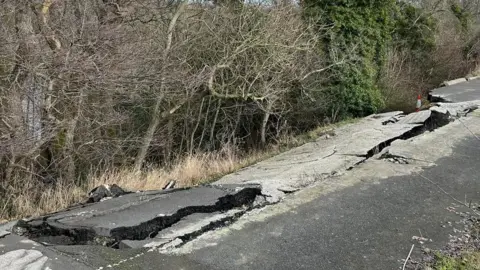 A road in very poor condition with half of it cracked off from the whole and sliding down the hill. A traffic cones can be seen at either side.