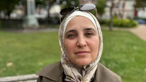 Tuka looks at the camera with a straight face. She is wearing a brown coat and a cream hijab.