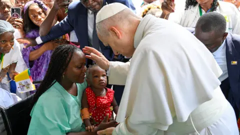 Reuters The Pope is blessing a woman in green, who is holding a toddler girl on her lap. There is a crowd of people around them