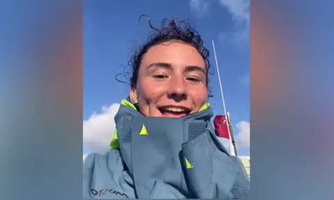 Sway PR Zara Lachlan, whose dark hair is pulled back from her face. It is a close-up of her sitting in a boat in the Atlantic. She is wearing a water-proof coat and a purple fleece can be seen beneath. Behind her can just be glimpsed blue ocean on her right and above is blue sky