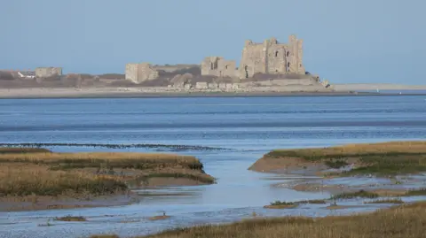 Timeandtides View of the ruined pale stone castle standing on Piel Island off Barrow-in-Furness. With blue water in front and a greyish blue sky behind. There are raised bits of land topped with grass, between which the sea finds a path.