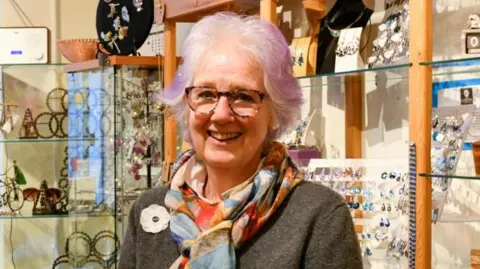 Rachael Rodway has silver and lilac short hair. She wears tortoise-shell glasses, a multi-coloured scarf and a grey wool jumper. She stands in front of a jewellery cabinet in a shop. 