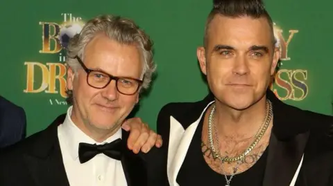 Getty Images A man with grey hair and glasses and a black bow tie standing next to Robbie Williams