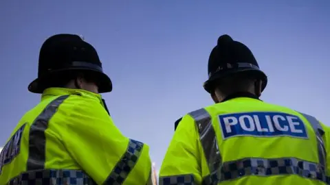 Two police officers in helmets and high-vis jackets stand with their backs turned to the camera under a blue sky.