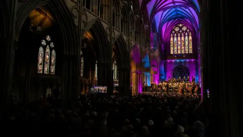 Lichfield Festival A crowd of people sit and watch a live orchestra performing in a dimly lit Lichfield Cathedral.