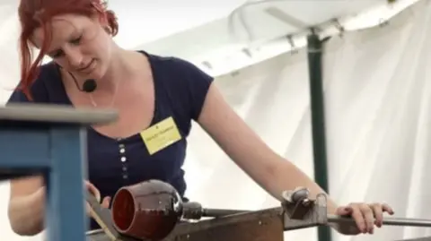 A young woman with red hair and a navy t-shirt is blowing glass to create a short brown pot. She has a gold name badge on.