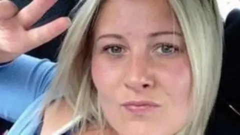 A woman with short blonde hair. She is wearing a blue top and pouting at the camera as she holds one hand up in a peace sign.