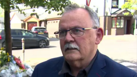 Maolíosa McHugh has short grey hair thinning on the top and glasses. He is wearing a navy blazer and brown shirt