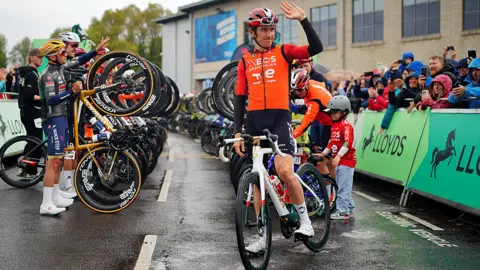 PA Media Professional cyclists form a line and hold their front tyre off the ground in a salute to Thomas, watched by fans