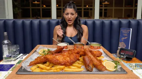 Leah Shutkever A woman sits in a restaurant with a large plate fish and chips on the table in front of her. She has straight dark hair and she has a mock shocked expression.