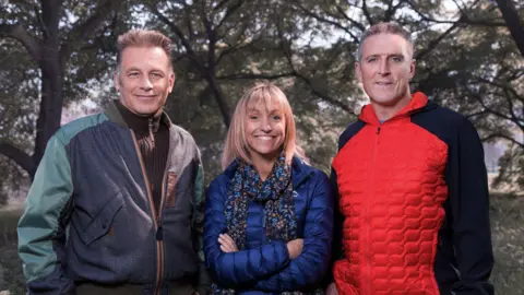 Jo Charlesworth/PA Wire The Winterwatch presenters are standing side by side in a professional picture. Mr Packham has short brown hair and is wearing a brown zip-up fleece, and a blue/ green jacket. Ms Strachan has short blonde hair with a fringe and is wearing a dark blue coat with a patterned scarf. Mr Williams has short dark hair and is wearing a red coat which has black sleeves.