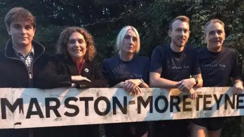 Nicola Haseler/BBC Five people line up behind a sign saying "Marston Moreteyne". Three are wearing running shorts and blue t-shirts with "CAPTAIN TOM" in white writing. The other two are wearing black fleeces and one is Hannah Ingram-Moore. There are trees in the background.