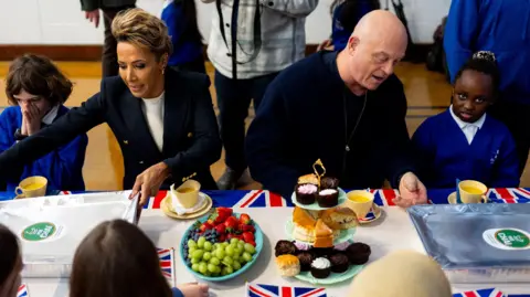 Dame Kelly Holmes and Ross Kemp sitting down at a table having lunch with school children dressed in blue, with decorative union flags in view.