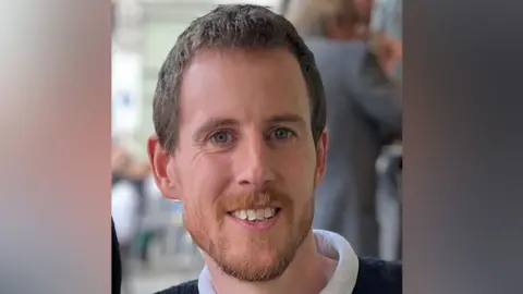 PC Miller with short brown hair and beard smiling at the camera. He is wearing a blue sweater with a white neck