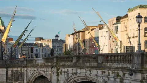 Getty Images A bridge over the River Lee, buildings are illuminated in sunlight on either side of the bridge. There are yellow flags on each street side. 