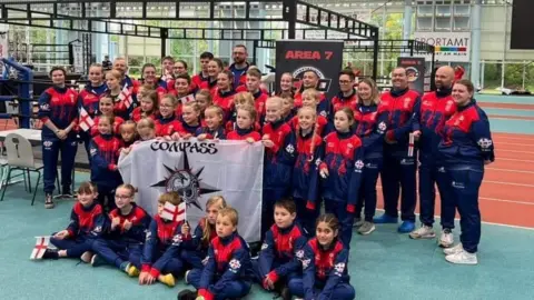 More than 45 competitors, including children, in red and blue training uniforms, pose holding a white Compass Martial Arts flag at the competition in Frankfurt, Germany.