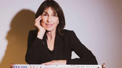 Sonica A woman with dark hair to her shoulders, wearing a black suit and standing behind a synthesizer keyboard.