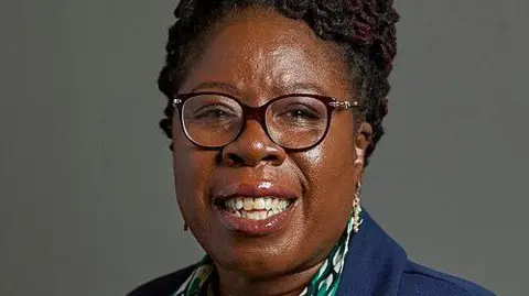 UK Parliament A posed shot in front of a grey background of a woman with black hair in black framed glasses. The collar of a navy jacket and black, green and white patterned shirt can just be seen.
