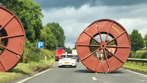 Two large brown cable reels on a motorway road in front of some cars.