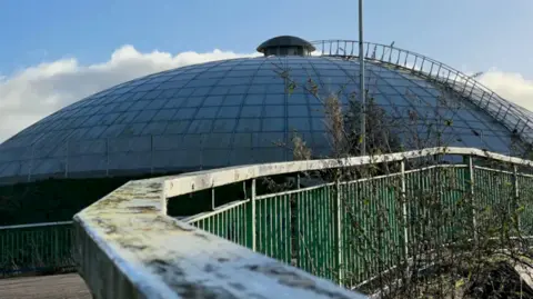 Aled Thomas The Oasis Leisure Centre exterior. There is a large dome with a ladder running to the top. There is a pavement with a green railing running across to the right.