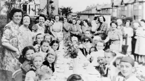 Getty Images A black and white photo of a street party in 1945. Children and women are crowded around a long table smiling at the camera.