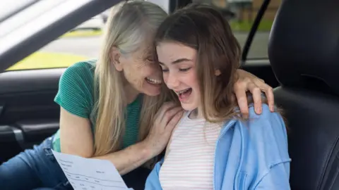 Side view of a young teenage girl opening her GCSE exam results in the passenger seat of the car. She is sitting next to her supportive mature mother, they are celebrating the good news, and they are both happy and smiling.