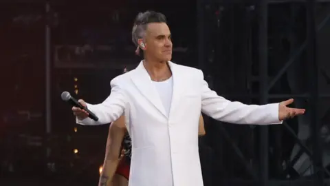 Robbie Williams is dressed in an all-white suit on a stage in front of thousands. There are backing dancers dressed in sparkly costumes behind him.
