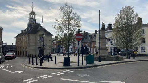 Brackley market square with trees and monument.  Vehicles parked along the street.