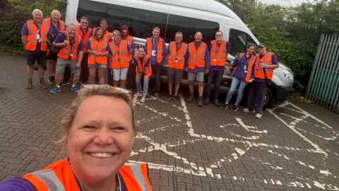 A woman takes a selfie with a group of people in hi-vis jackets assembled in front of a van in the background.