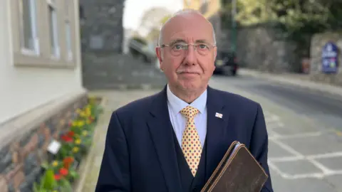 An older man wearing a navy suit, pale blue shirt and a yellow tie with a red and blue diamond pattern. He wears gasses, has very short grey hair, and is standing outside Guernsey's royal court. 