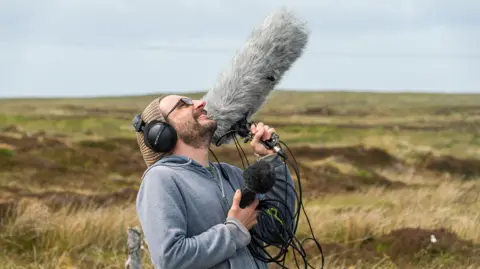 Hopscotch A man with a beard and a brown beanie hat in a grey hoodie holds a large microphone in the air while out in a rugged Scottish landscape