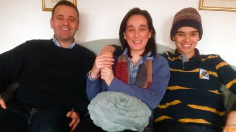 Ballester family Pedro Ballester has a black rugby shirt with yellow horizontal stripes and a brown beanie hat. His father Pedro has a black hair, black sweater and sky blue shirt. His mother Esperanza has long brown hair, a blue cardigan, red and breown scarf