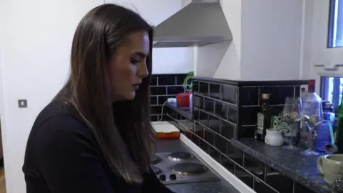 Hannah is in her kitchen washing dishes wearing a black jumper