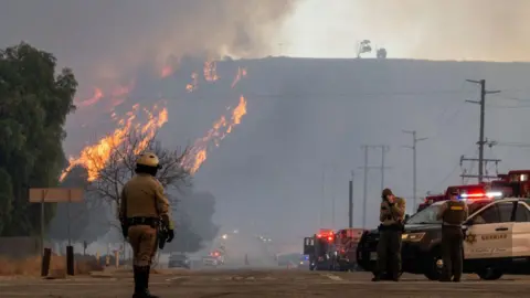 Getty Images Emergency workers patrol the street as flames burn and smoke billows into the air in the distance