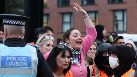 Reuters One protester, wearing pink, holds her arm up in the air and shouts as a police liaison officer stands to the left and crowds surround the woman
