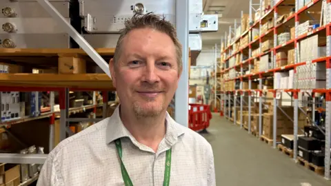 Martin Heath/BBC A large hall with a grey floor and wood shelving in a metal frame. The shelves are full of brown and white boxes of various sizes. Stuart Turpin, with short brown hair and a slight beard, wearing a white shirt and green lanyard, is in the foreground.