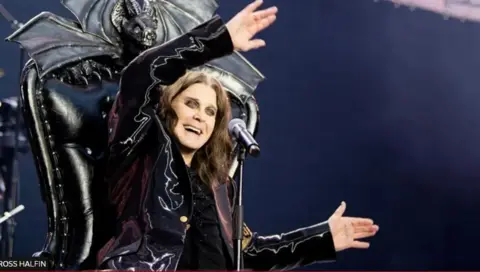 A long-haired man sits in a throne with a black bat design behind him. He is wearing a purple-ish velvety looking coat, is waving his arms and smiling. He has thick black eyeliner.