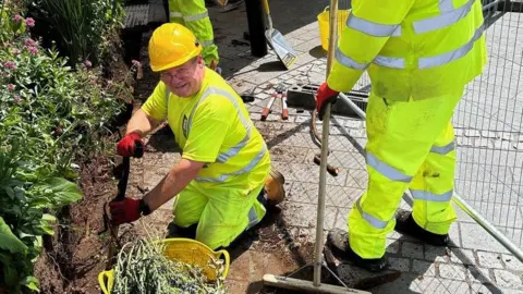 A man on his knees, wearing high visibility clothing, a yellow hard hat, glasses and red gloves. There is a yellow bucket in front of him with weeds. To his right is another man in high visibility clothing. There is concrete underneath him. Behind is a temporary metal fence. 