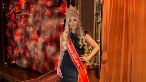 Emily Kathleen The image shows a blonde woman standing in a theatre box, wearing a crown and a bright red sash that reads "Ms Glamour UK 2025".