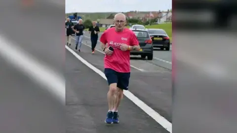 Kelly Russell A man with white shaved hair running at the side of a road with cars travelling down the side. He's wearing a pink Blaydon Races T-shirt and holding his phone. People can be seen running behind him.