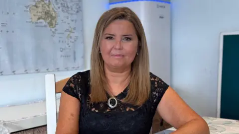 BBC/Richard Moss Dr Rae Duncan has long blonde/brown hair, a black top and a silver necklace with oval pendent. She is sat on a chair in a room with a global map on the wall.