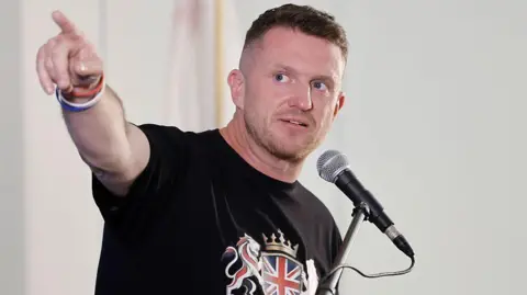 Far-right activist Tommy Robinson, wearing a black T-shirt with a union jack emblem, speaking into a microphone and pointing to his right.