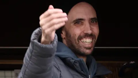 Tal Shoham, dressed in grey anorak, blue hoodie and black hat, smiles and gestures towards a crowd after being released by Hamas. 