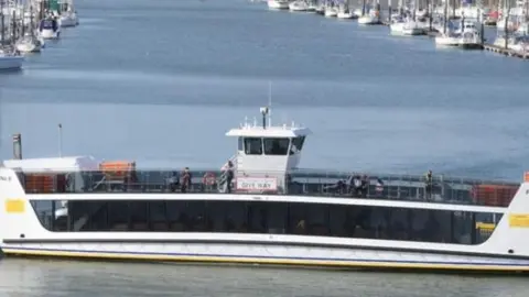 The Floating Bridge 6 floats in a marina. It is white, with large blacked out glass panelling and there are multiple boats in the background also docked in the marina