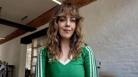 Kathy, who is has long curly brown hair with a fringe, wearing a green V-neck Adidas top