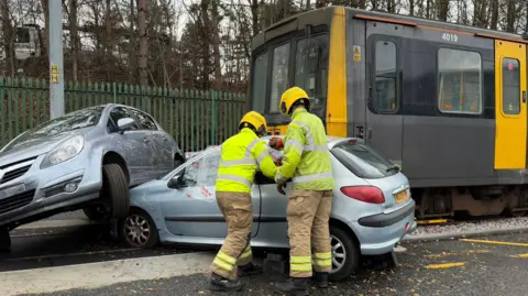 BBC A training exercise that makes it look like a grey and yellow metro train has hit two grey cars at a level crossing. Two firefighters, who are wearing bright yellow helmets, are working on the nearest car.