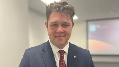 Owen Sennitt/BBC Man with brown hair in a blue suit smiles to camera. 