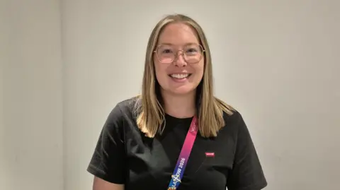 BBC Alice McNaugher - a woman with blond hair past her shoulders, glasses and a black top, wearing a pink coloured lanyard. She is standing inside a room, smiling for the camera. 