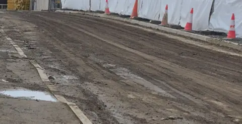 Jackie Gardner Another road not covered by tarmac and instead is muddy with tyre tracks across it. On the right side of the road there are orange cones in front of a white fenced off area.